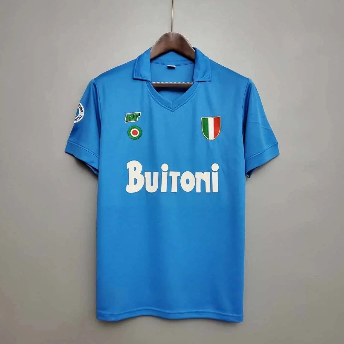 Blue sports jersey with 'Buitoni' branding on a hanger against a grey background. The shirt is a 1987 Napoli home kit