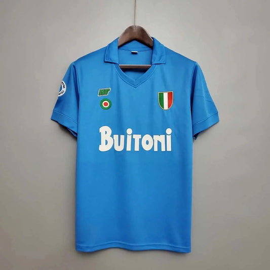 Blue sports jersey with 'Buitoni' branding on a hanger against a grey background. The shirt is a 1987 Napoli home kit